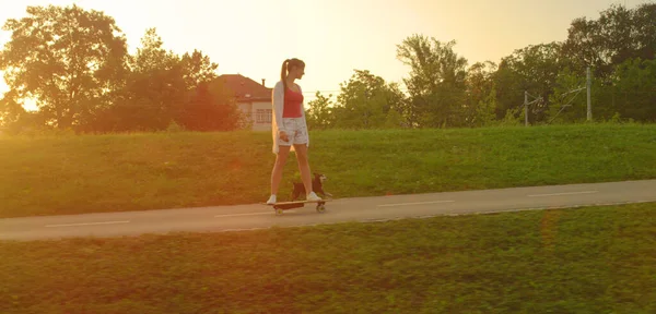 LENS FLARE: Glücklicher kleiner Hund läuft neben Mädchen auf Elektro-Skateboard. — Stockfoto