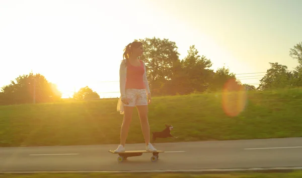 ZON FLARE: Jonge vrouw rijdt op een elektrisch skateboard terwijl haar puppy loopt langs. — Stockfoto
