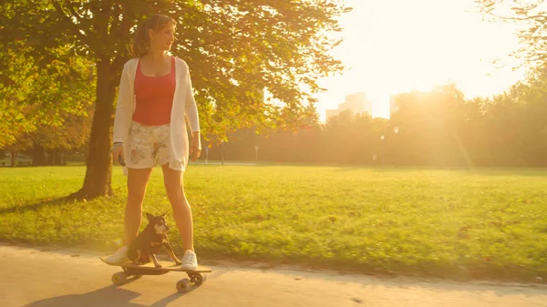 SUN FLARE: boldog fiatal nő és idősebb kutya lovaglás egy elektromos longboard. — Stock Fotó