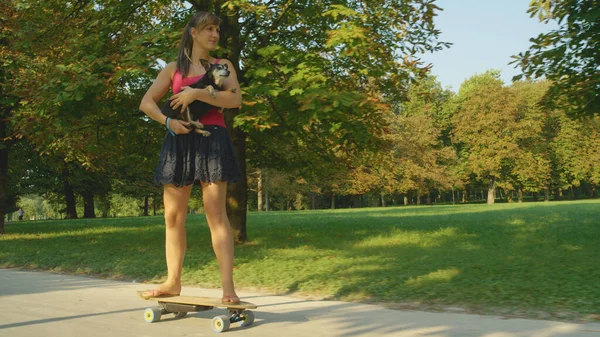 Cute shot of woman holding her puppy in her lap while skateboarding through park — Foto de Stock