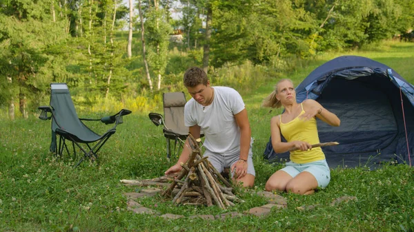 Junge Frau versucht Insekten abzuwehren, während sie mit Freund das Lagerfeuer zubereitet. — Stockfoto