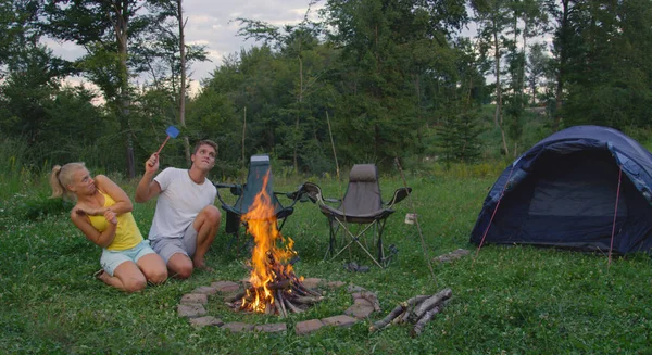 Caucasian man and woman sitting by campfire waving a fly swatter to repel bugs. —  Fotos de Stock