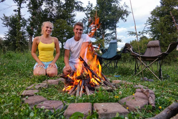 PORTRAIT: Casal alegre em férias de acampamento ajoelhado perto da fogueira quente. — Fotografia de Stock