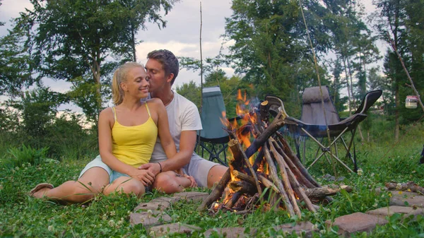 CLOSE UP: Happy man kisses his girlfriend on cheek while sitting by a campfire. — Stock Photo, Image