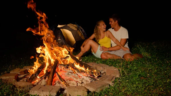 CLOSE UP: Cheerful young Caucasian couple sits and laughs by the warm campfire. — Stock Photo, Image