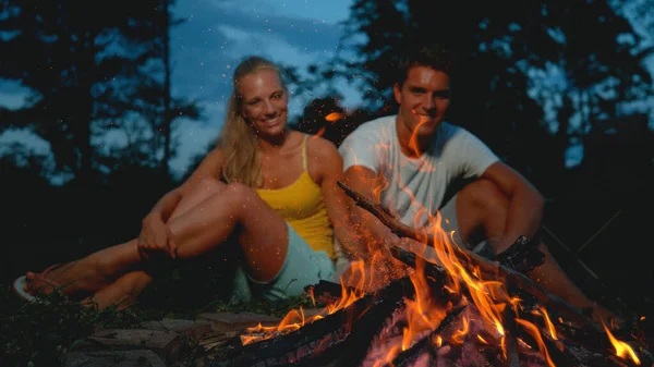 CLOSE UP: Cheerful man pokes fire with a stick while camping with girlfriend. — Stok fotoğraf