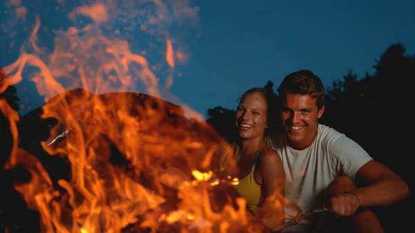 CLOSE UP: Joyful man pokes the campfire with a branch while cuddling with girl — Stok fotoğraf