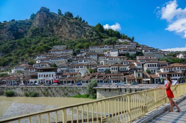 Female tourist in red dress stands on bridge and observes the city of Berat. clipart