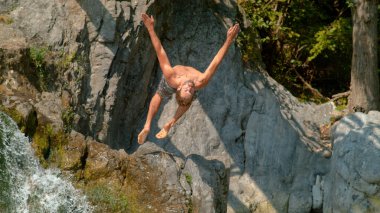 Athletic Caucasian guy does a cool backflip off the edge of a tall waterfall.