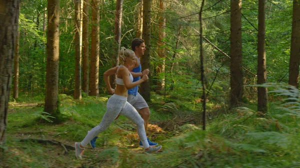 Atlétikai kaukázusi férfi és sportos barátnője kocognak a napsütötte erdei ösvényen. — Stock Fotó
