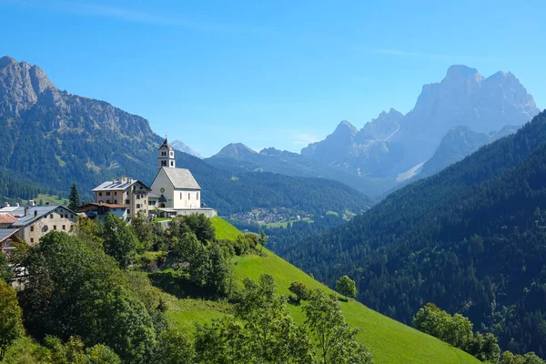 AERIAL: Malá vesnice na vrcholu zeleného kopce, obklopená italskými Dolomity. — Stock fotografie