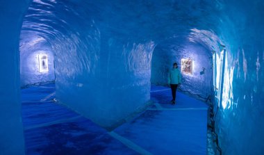 Female tourist walks through the breathtaking blue lit gallery inside ice cave. clipart