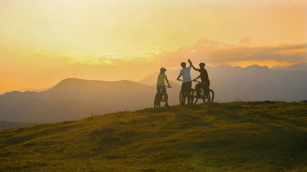 Üç arkadaş bisikletlerini bir tepenin kenarına sürüp gün batımında beşlik çakıyorlar.. — Stok fotoğraf