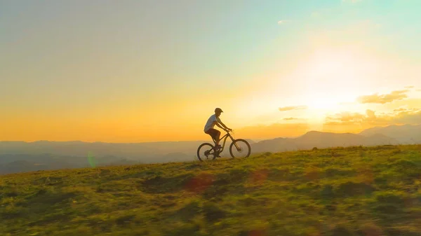 DRONE: Athletic tourist rides an electric bicycle up a grassy hill at sunrise — Stok fotoğraf
