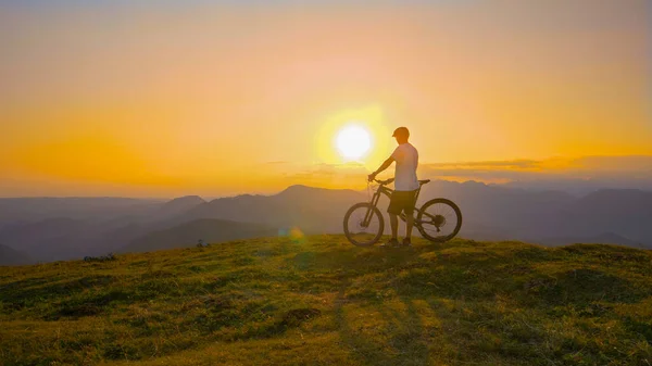 Bisiklet sürücüsü çimenli bir tepeden güneşin doğuşunu izliyor.. — Stok fotoğraf