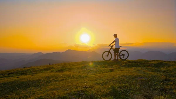 Kır bisikletçisi bir tepenin üstünde durur ve gün batımını izler.. — Stok fotoğraf