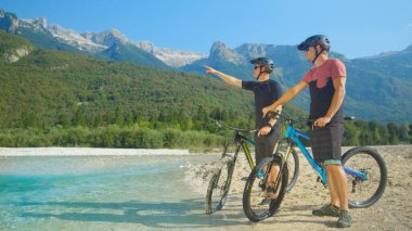Genç motorcular turkuaz nehrinin kenarında dinleniyor ve güzel dağları gözlemliyorlar..