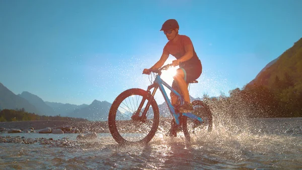 LOW ANGLE: Fit man jeździ na rowerze elektrycznym w płytkiej rzece i chlapie wodą. — Zdjęcie stockowe