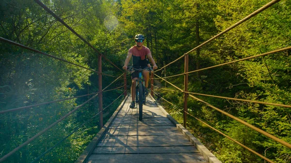 FLARE DE LA LENTE: Hombre alegre monta una bicicleta a través de un puente colgante en un día soleado. —  Fotos de Stock