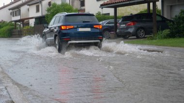 Mavi SUV bir banliyö mahallesinin sular altında kalmış sokaklarından geçiyor.