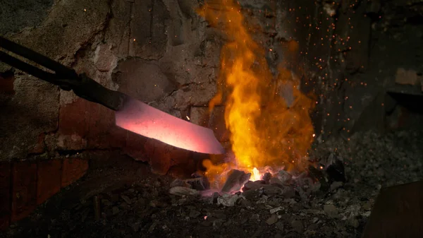 FECHAR-SE: Ferreiro puxa uma lâmina de faca para fora do fogo que arde no forno. — Fotografia de Stock