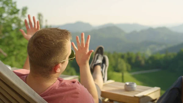 CLOSE UP: Man waves his hands while lying in lounge chair and talking to friend — Stock Photo, Image