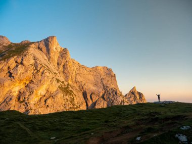 Alplerdeki çimenli tepenin kenarında duran yürüyüşçü zaferle kollarını kaldırdı.