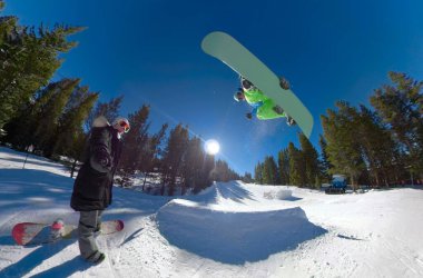 Snowboard sporcusu vurucudan atlayıp bir numara yaparken kadın selfie çekiyor.