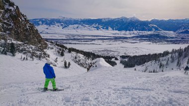 Snowboardcu bir kayak merkezi yamacından aşağı inmeden önce karlı araziyi gözlemler.