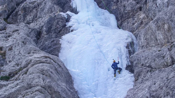 Αθλητικός άνθρωπος με κραμπόνια και τσεκούρια πάγου σκαρφαλώνει στον πανέμορφο παγωμένο καταρράκτη. — Φωτογραφία Αρχείου