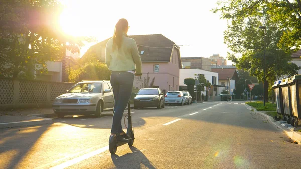 FLARE DE LA LENTE: Mujer joven monta un scooter eléctrico por un camino vacío en los suburbios — Foto de Stock