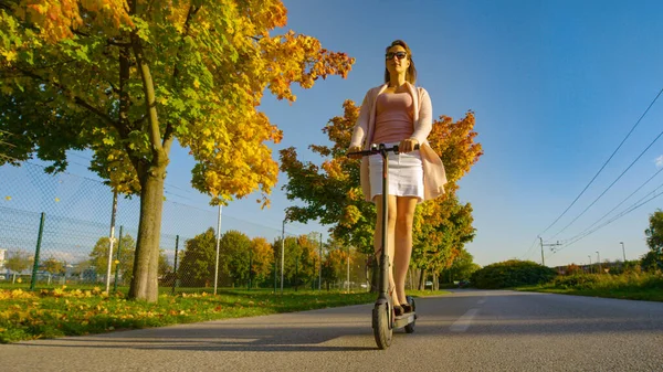 ÁNGULO BAJO: Chica sonriente en falda blanca y tacones altos monta un scooter eléctrico. —  Fotos de Stock