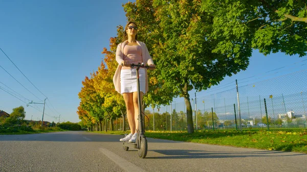 LOW ANGLE: Jonge vrouw pendelt langs een val gekleurde laan op een e-scooter. — Stockfoto