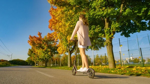 ÁNGULO BAJO: Mujer moderna montando un scooter eléctrico a través de los suburbios en otoño — Foto de Stock
