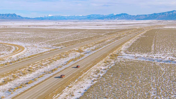 DRONE: Vliegen langs een asfaltweg leidt voertuigen over de besneeuwde woestijn. — Stockfoto