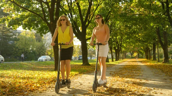 Két boldog női barát beszél, miközben robogóznak az őszi színes parkban. — Stock Fotó