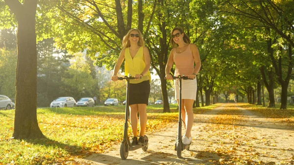 Golden autumn sunbeams shine on cheerful women riding e-scooters around the park — Φωτογραφία Αρχείου