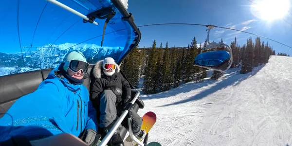 Sluiten: Snowboarder paar glimlacht tijdens het rijden op de stoeltjeslift op een zonnige dag. — Stockfoto