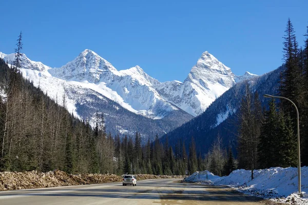 Βρώμικο λευκό SUV οδηγεί κατά μήκος μιας γραφικής εθνικής οδού που τρέχει κάτω από τα χιονισμένα βουνά — Φωτογραφία Αρχείου
