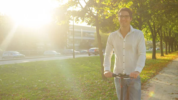 PORTRAIT: Jongeman glimlacht terwijl hij over een e-scooter in het midden van een laan staat Stockfoto