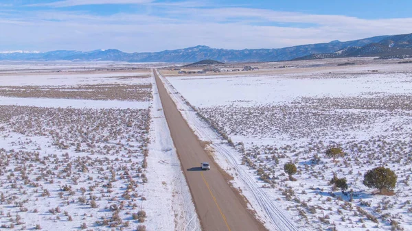 AERIAL: Scenic asfalt snelweg leidt zilveren SUV door het besneeuwde landschap. — Stockfoto