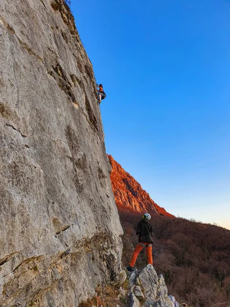 VERTICAL: Man looks up at female friend climbing up a rocky wall at sunset. — Stock Photo, Image
