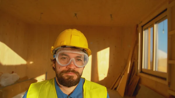 PORTRAIT Adam, kasketli ve güvenlik gözlüklü modern bir ahşap evde duruyor. — Stok fotoğraf