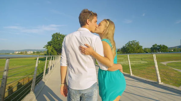 Twee jonge geliefden kussen elkaar terwijl ze elkaars hand vasthouden en een brug oversteken. — Stockfoto