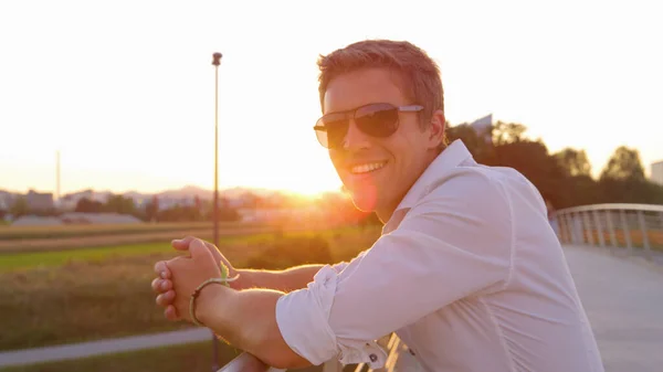 CERRAR: Joven hombre de negocios con gafas de sol sonríe para la cámara al atardecer —  Fotos de Stock