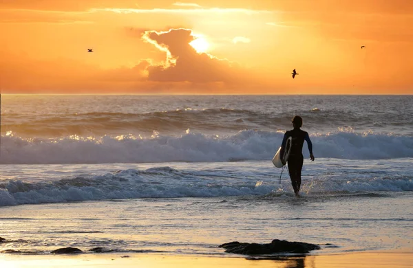 認識できない男サーファーは日没時に波に乗るために海に向かって歩く. — ストック写真