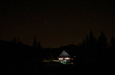 Yıldızlı gece gökyüzü, ormanın ortasındaki bir kulübenin üzerinde ışıklar açıkken uzanıyor.