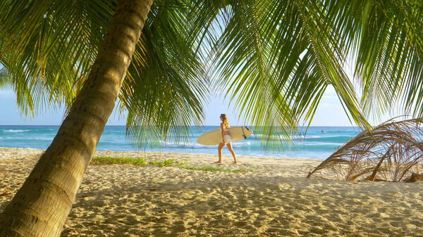 Young female tourist carries her surfboard along an idyllic tropical beach.