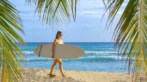 Femmina surfista porta la sua tavola da surf lungo la spiaggia tropicale la sera di sole — Foto Stock