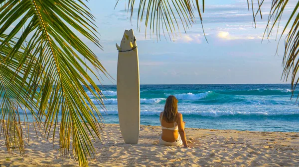 Jonge vrouwelijke surfer zit naast haar surfplank vast in het zand. — Stockfoto